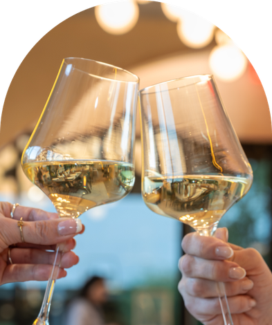 Two hands toast with glasses of white wine against a blurred, warm-lit background.
