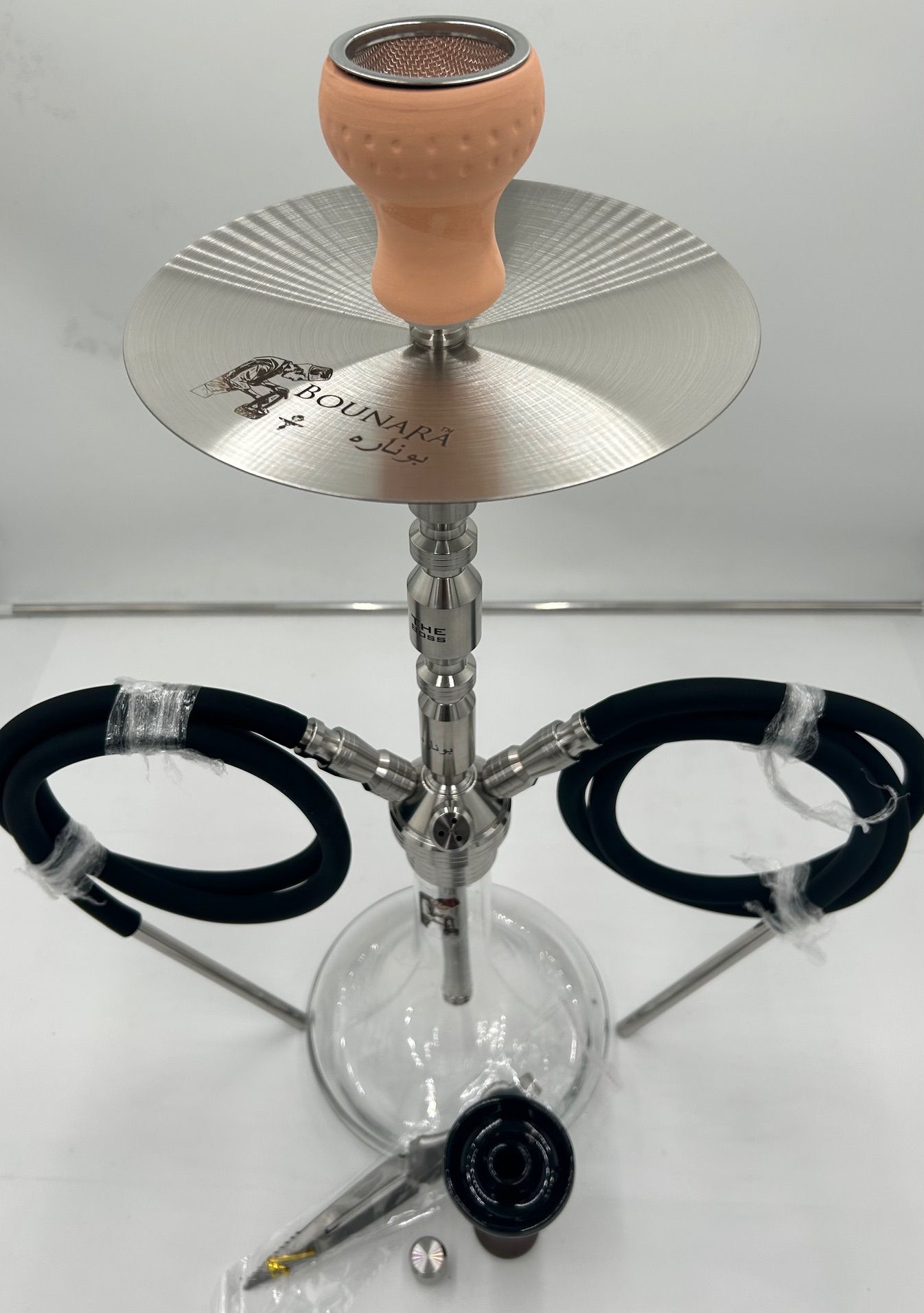 A modern silver hookah with a glass base, two black hoses, and a tan bowl, on a white surface.