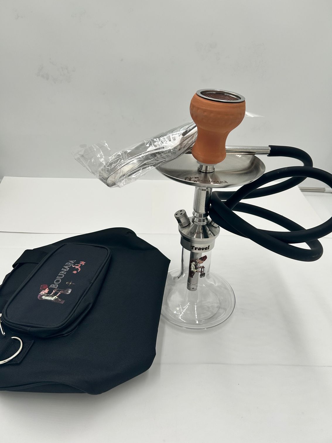 A hookah with a black hose, carrying bag, and ceramic bowl on a white surface.