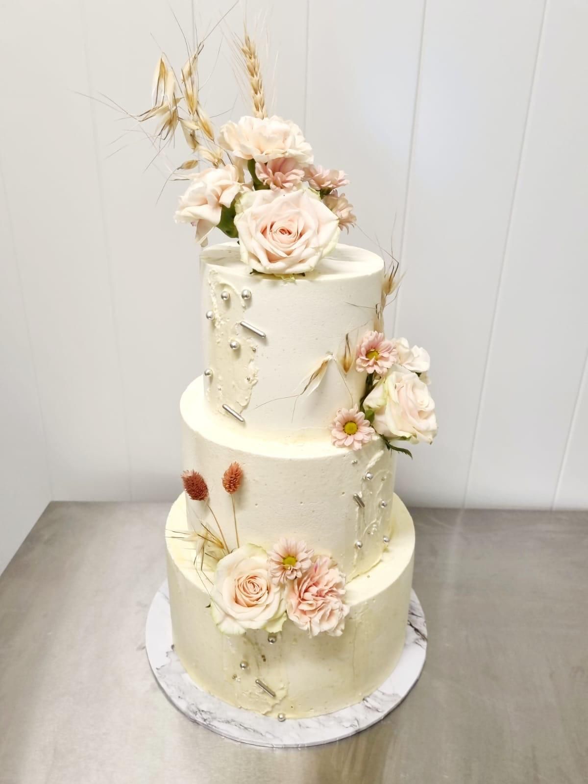 Dreistöckige Hochzeitstorte mit strukturierter weißer Glasur, verziert mit rosa Blüten und Weizenähren.