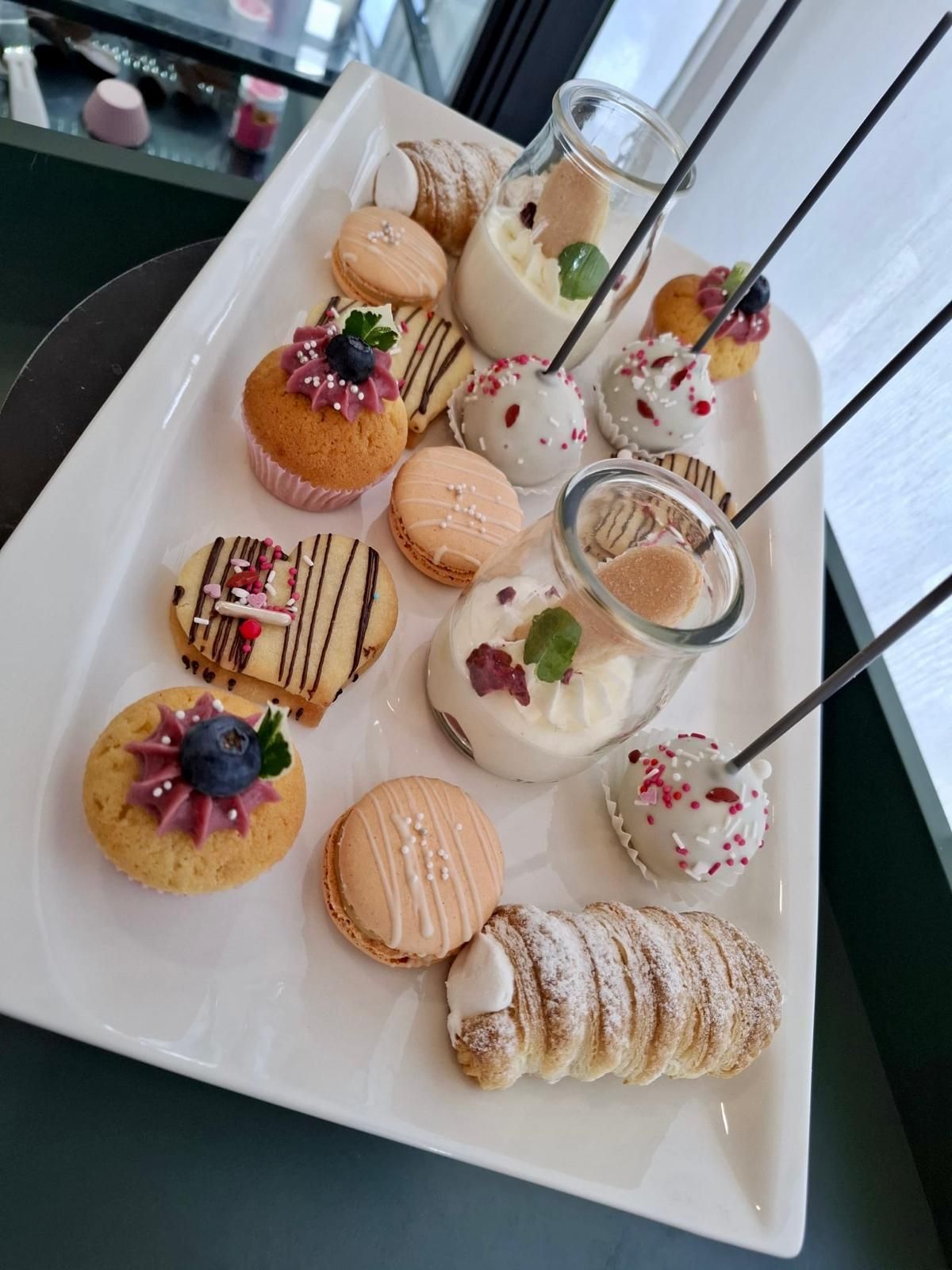 Auf einem Tisch steht eine Platte mit verschiedenen Desserts: Cupcakes, Macarons, Cannoli und Leckereien am Spieß.