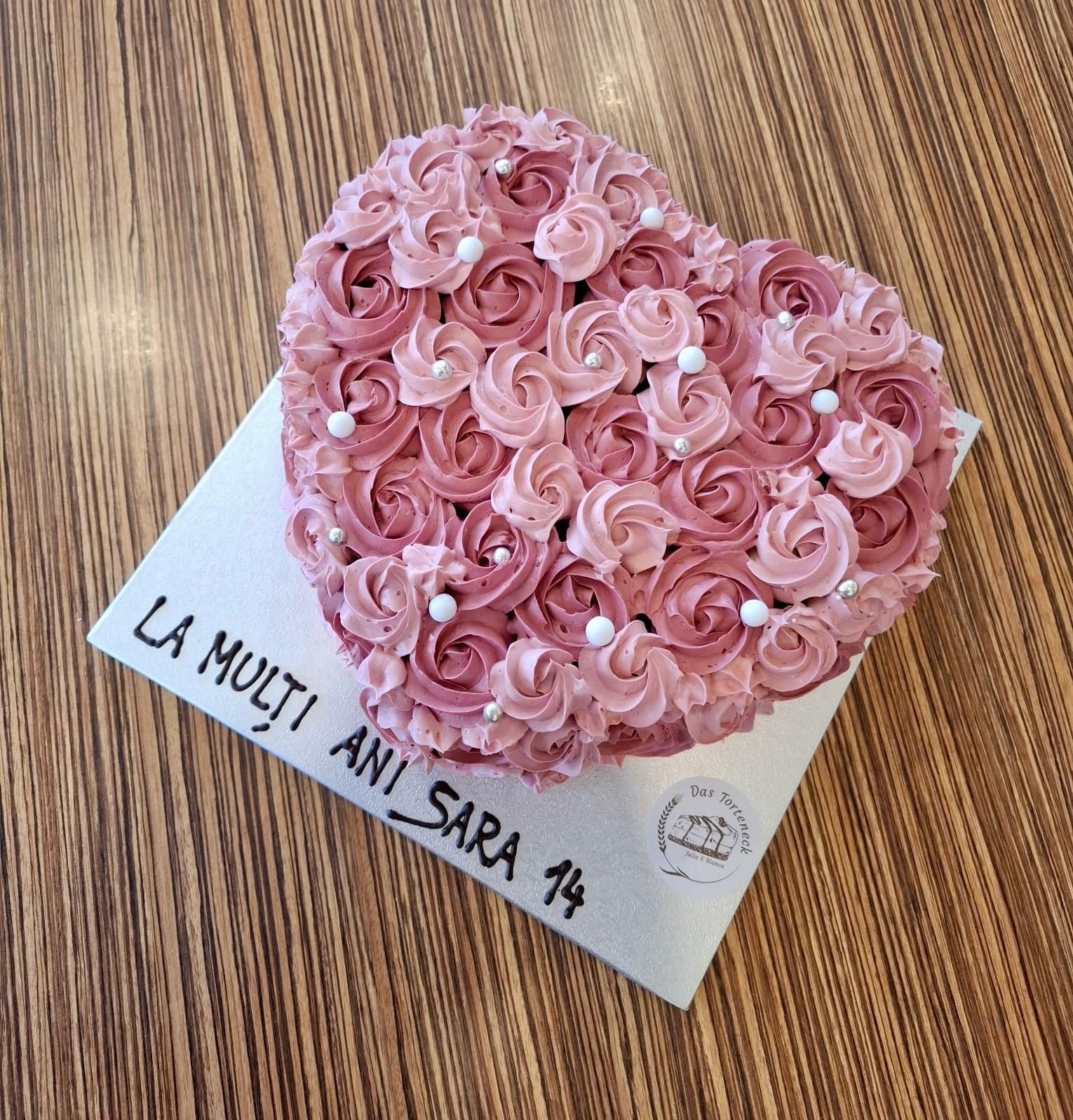 Herzförmiger Kuchen mit rosa Zuckergussrosen auf einem weißen Quadrat mit der Aufschrift „LA MULȚI ANI SARA 14“ auf einem Holztisch.