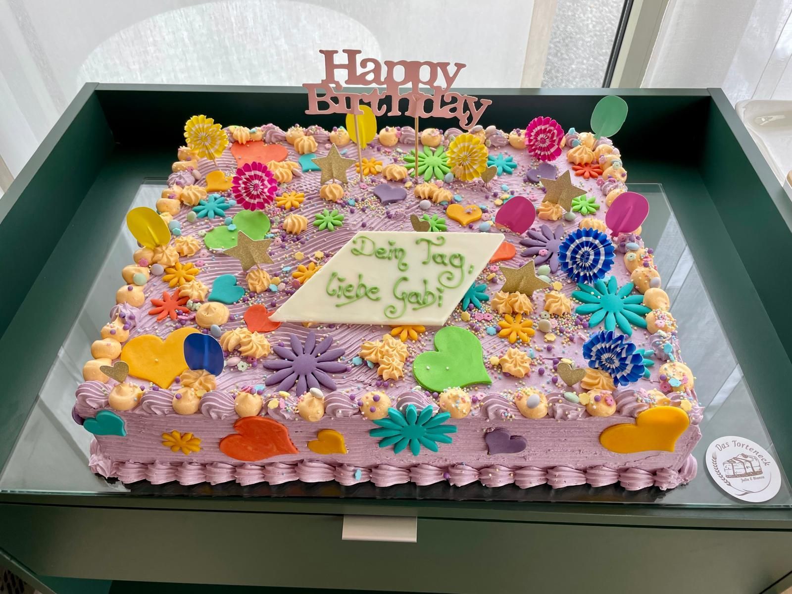 Quadratische Geburtstagstorte mit lila Zuckerguss, bunter Dekoration und einem „Happy Birthday“-Schriftzug.