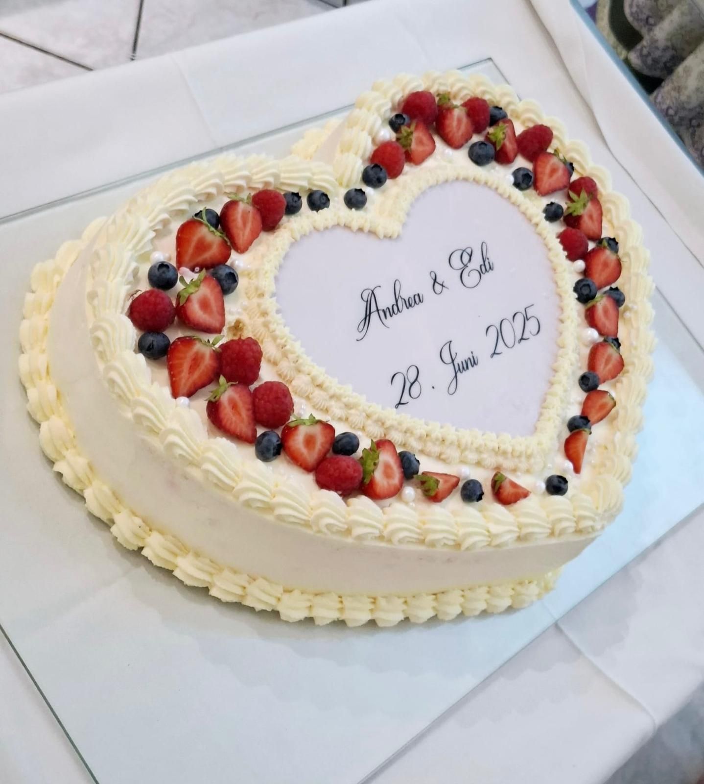 Herzförmiger Kuchen mit weißem Zuckerguss, verziert mit Beeren. Text: „Andrea & Erik 12. Juni 2015“.