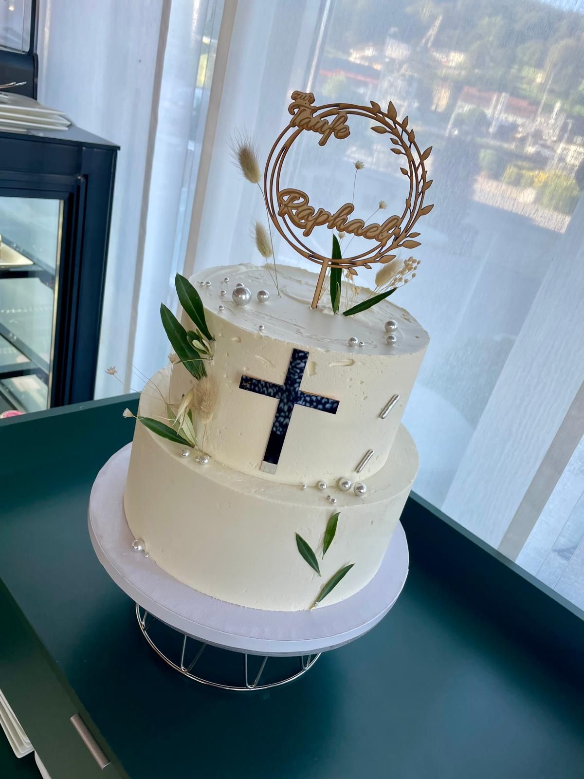 Zweistöckige weiße Torte mit Kreuz, Grünzeug und Namensschild auf einer Tortenplatte.