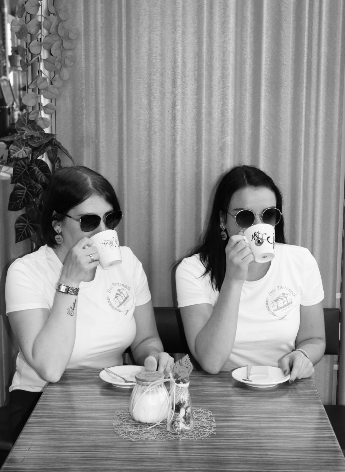 Zwei Personen mit Sonnenbrillen sitzen an einem Tisch und trinken aus Bechern.