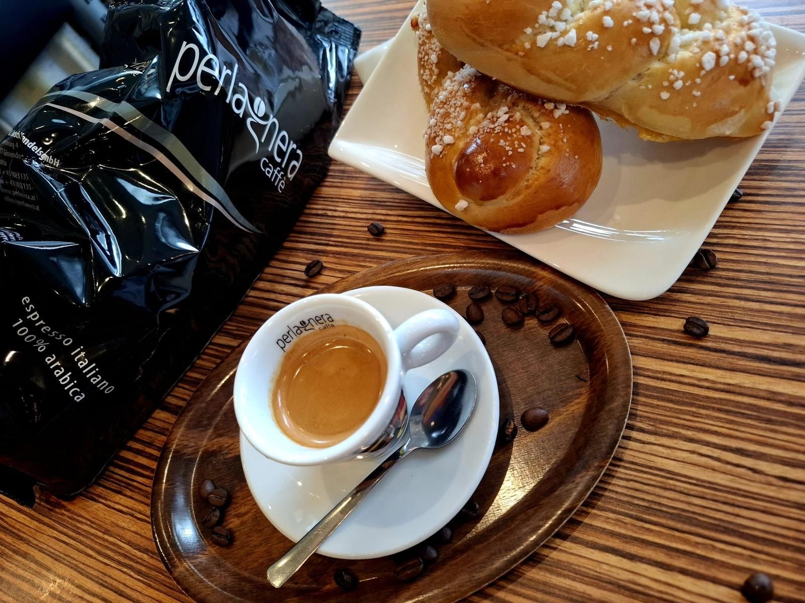 Espresso in einer Tasse mit Löffel auf einem Holztablett, dazu eine Tüte Kaffee und Brot.