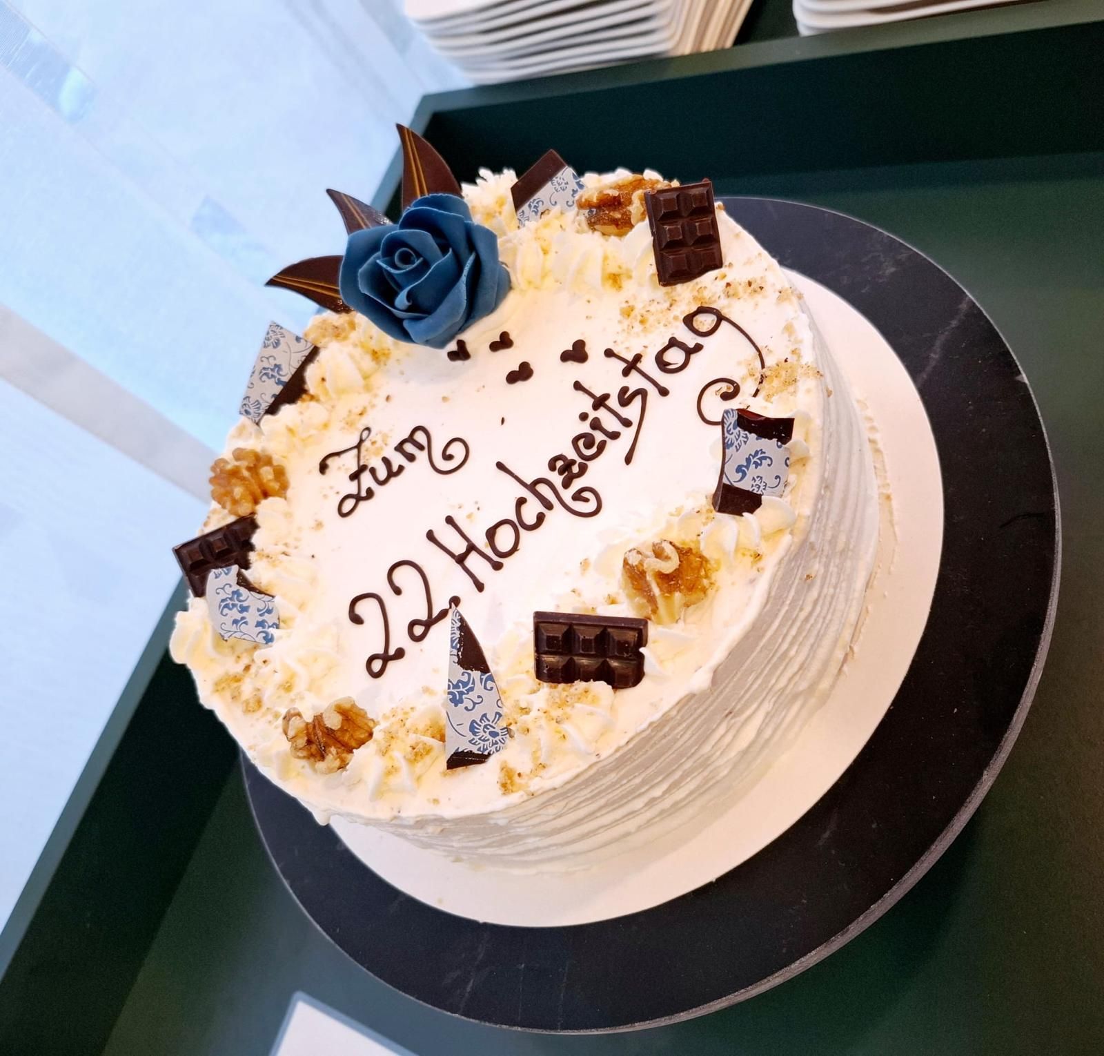 Kuchen mit weißem Zuckerguss und der Aufschrift „Zum 22 Hochzeitstag“, verziert mit Nüssen, Schokolade und einer blauen Blume.