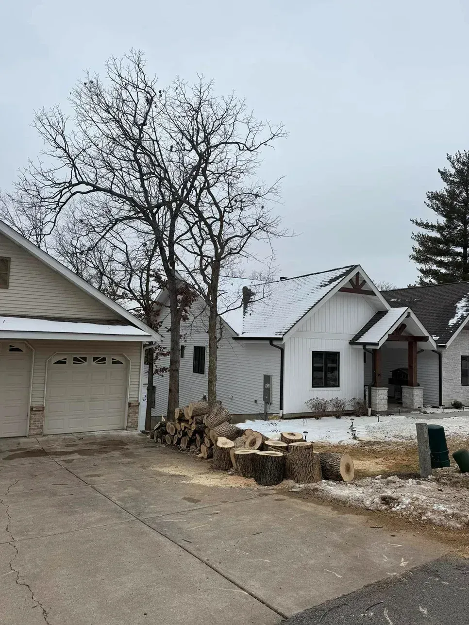 White houses and garage with snow-covered roofs and trees. Cut logs lie in the driveway.