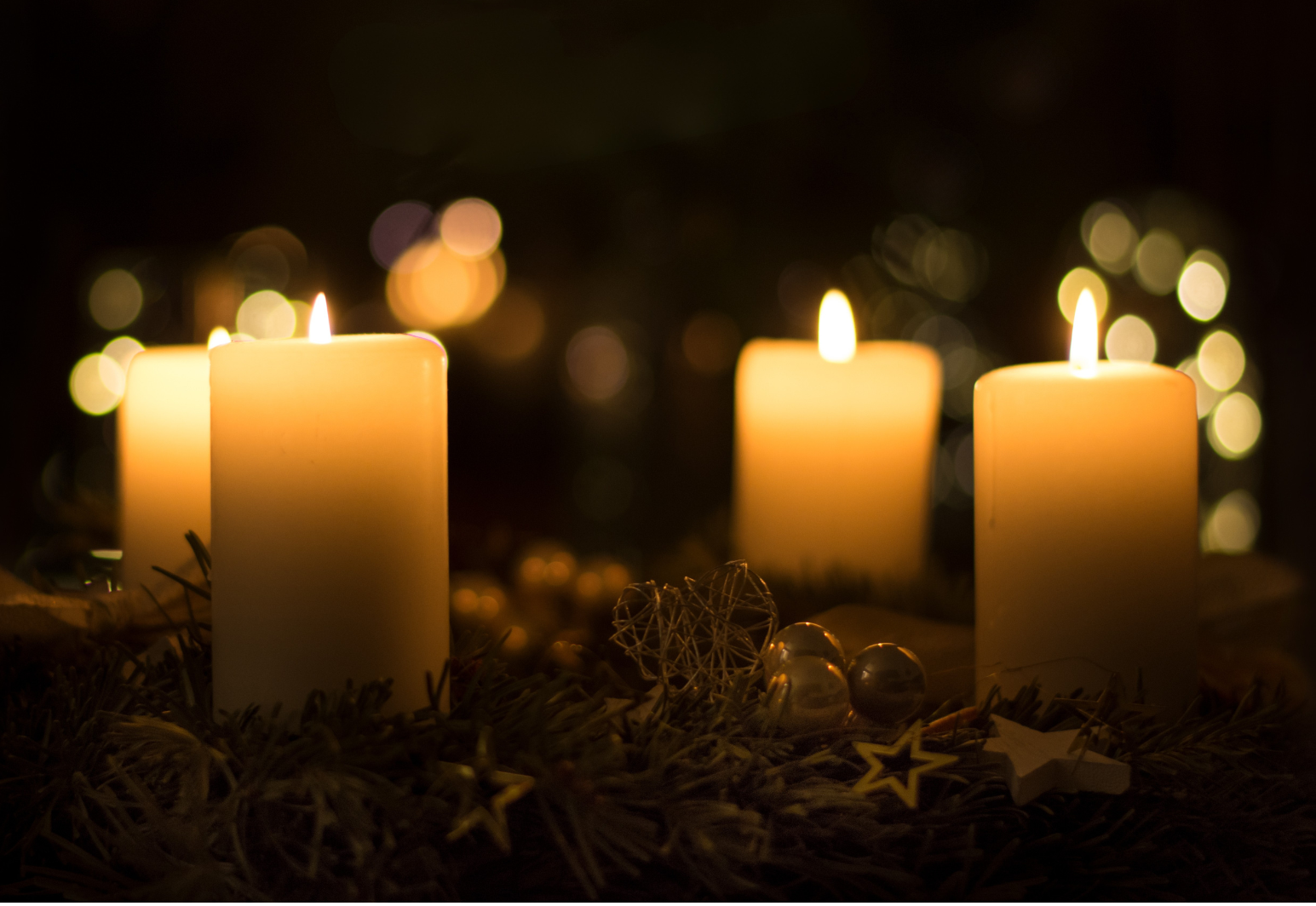 Four lit candles on a wreath with blurred lights in the background.