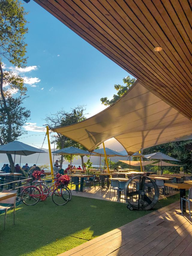 Pampa restaurante coatepeque al aire libre con sombra, mesas, bicicletas y toldos blancos junto a un césped soleado y un cielo azul.
