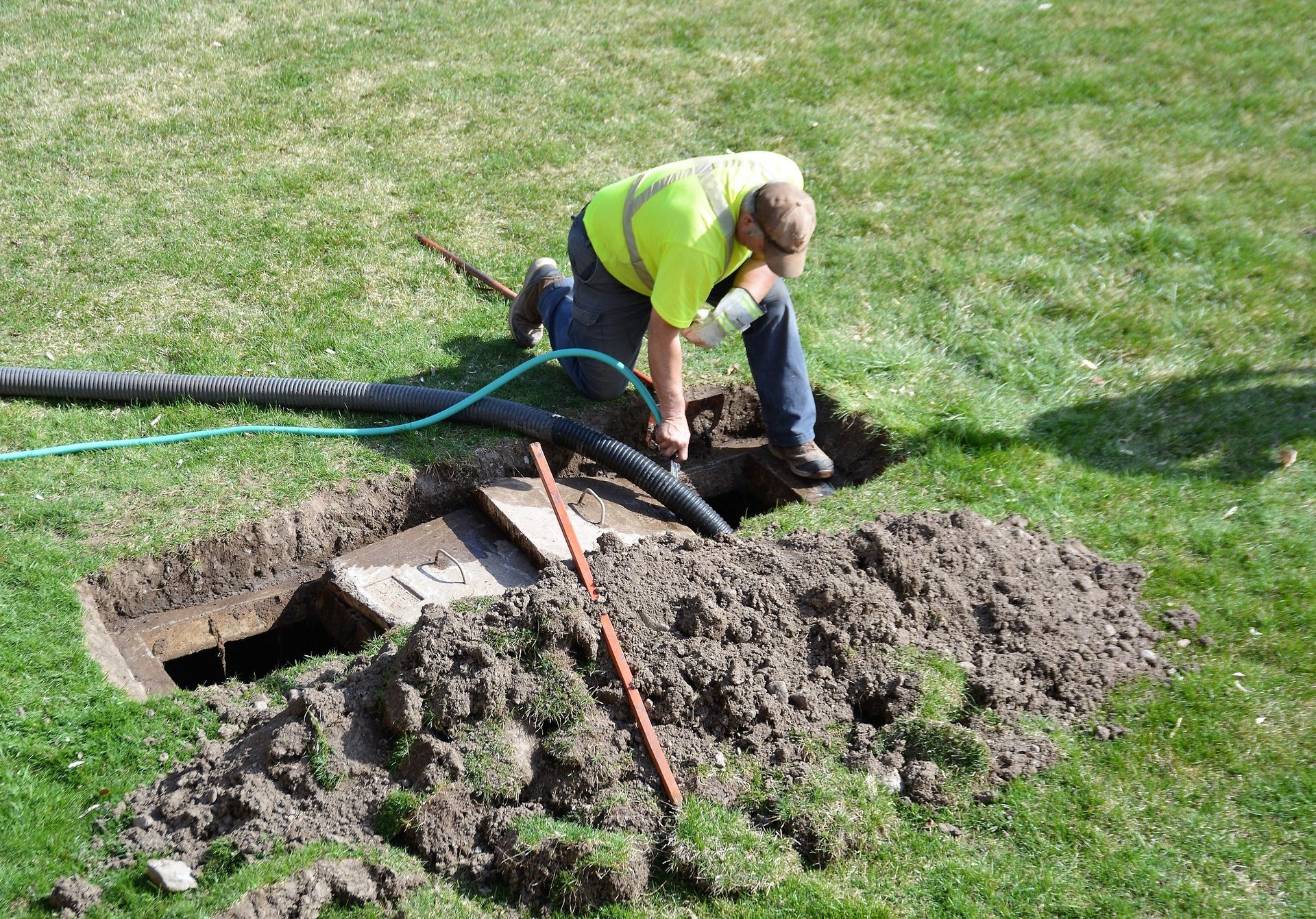 Worker in a neon vest digging soil beside an open trench and hose on a grassy lawn