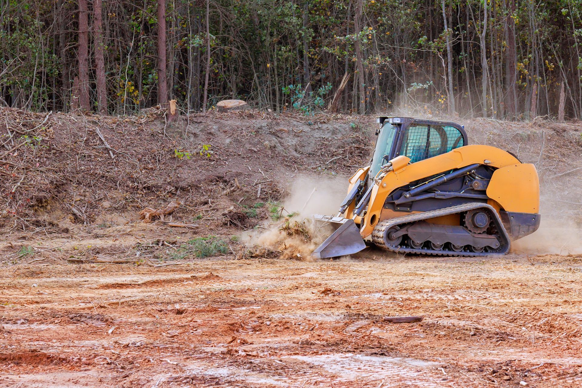 Orange skid-steer loader digging dirt on a construction site, kicking up dust near a wooded area
