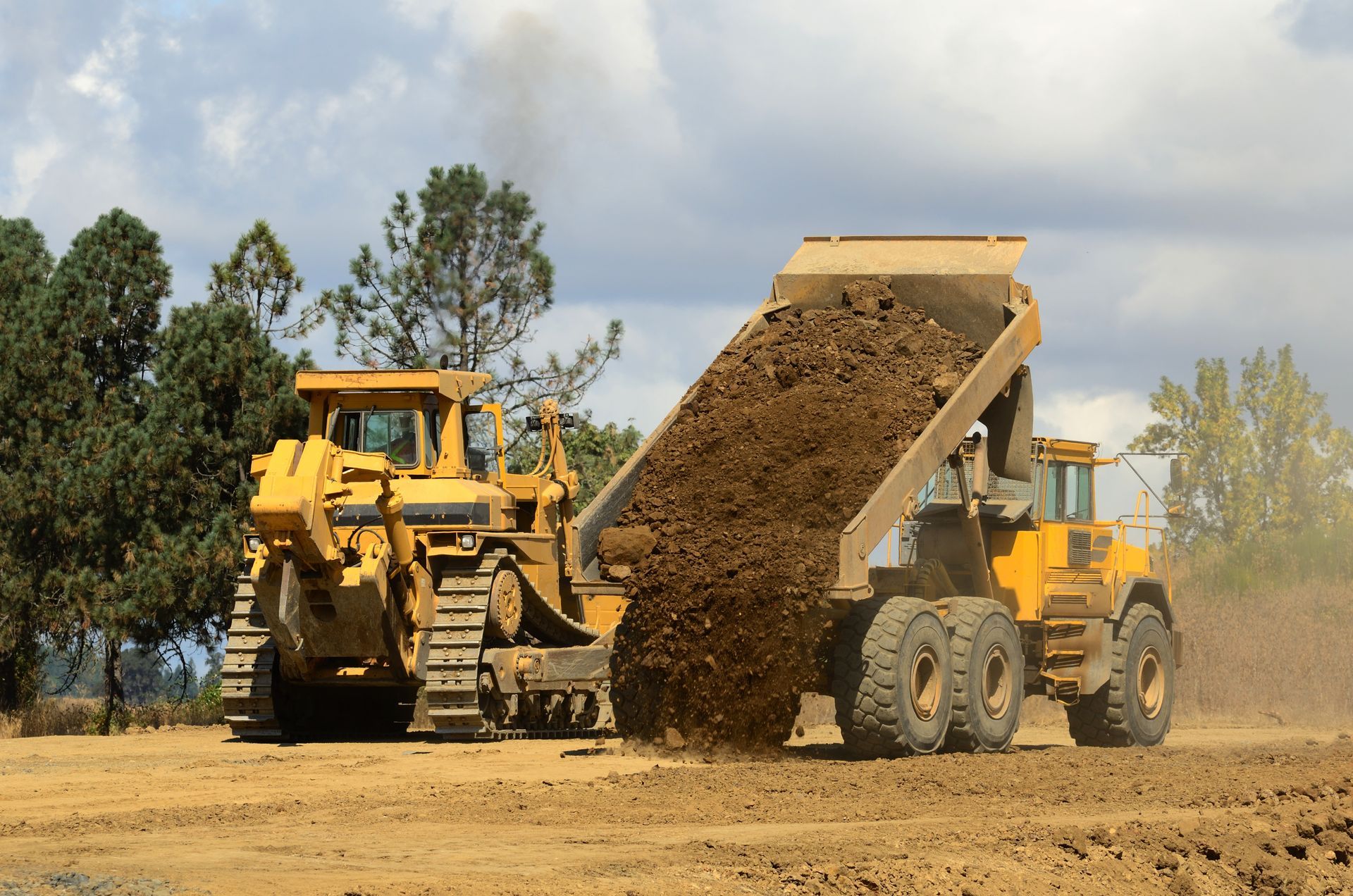 Two yellow dump trucks unloading dirt at a construction site with trees in the background