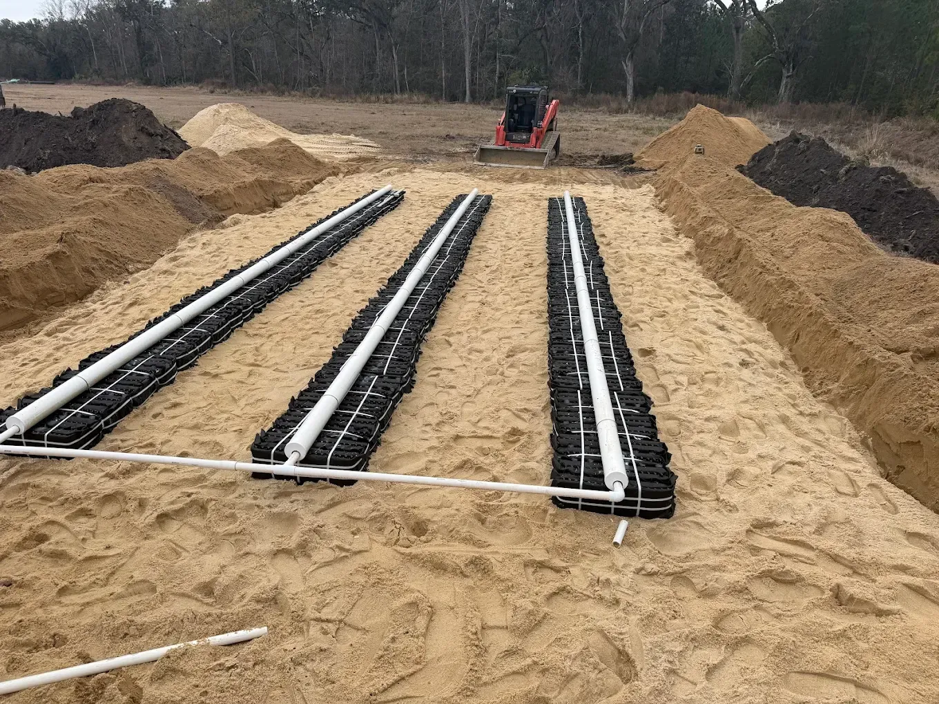 Three black drainage pipes laid in a sandy trench with a small machine in the background
