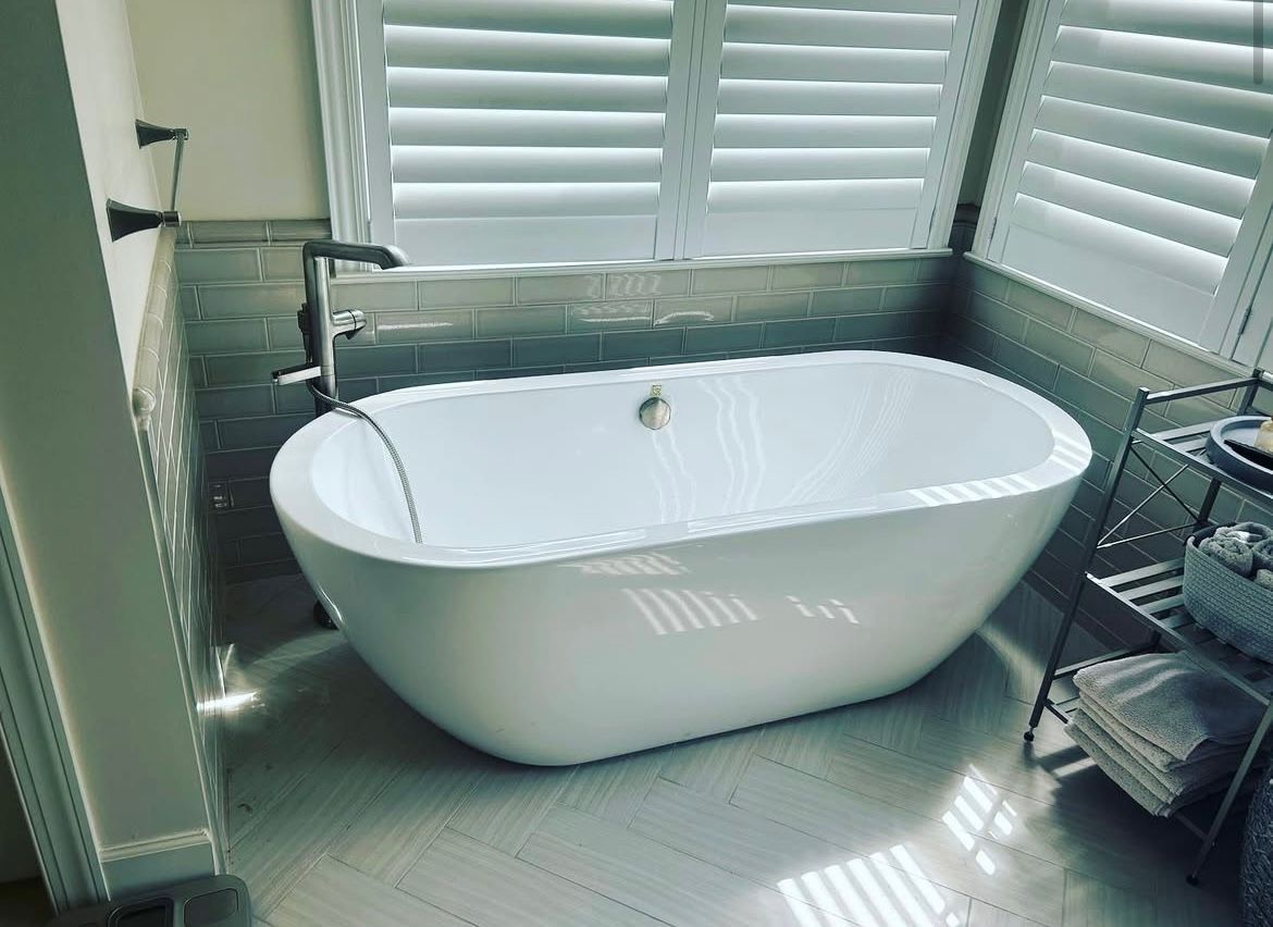 Modern bathroom with a white freestanding bathtub in front of a window with white shutters.