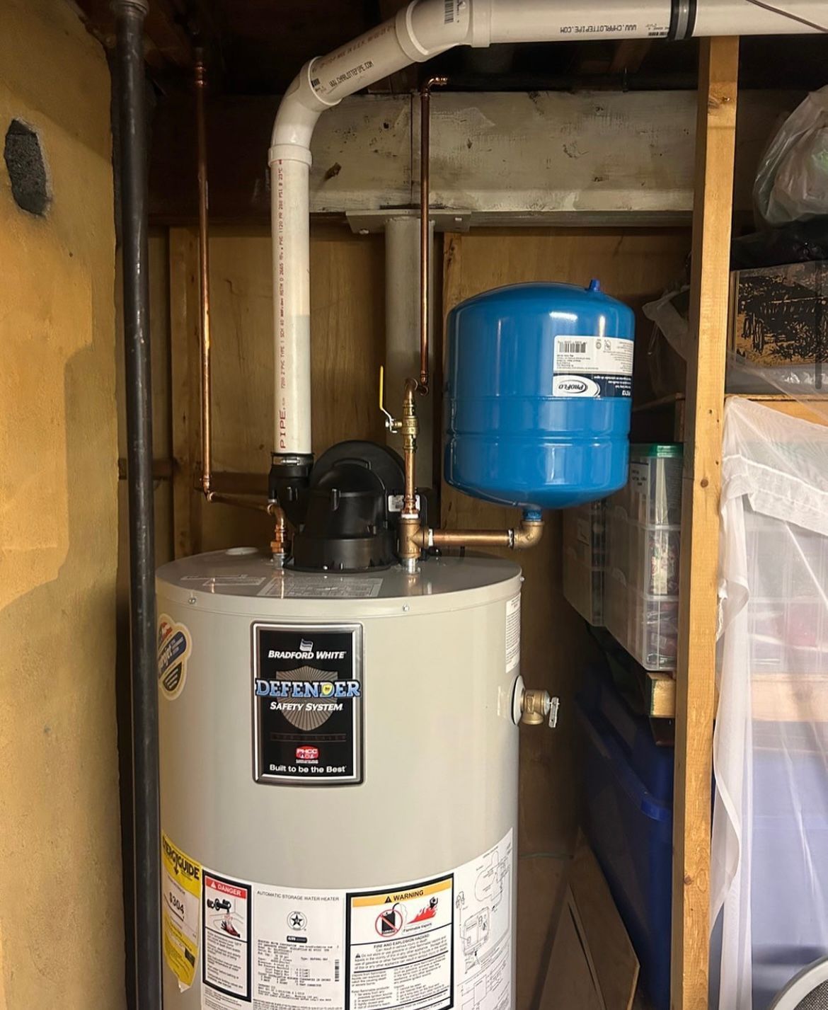 A hot water heater with white and copper pipes, plus a blue expansion tank, in a basement alcove.
