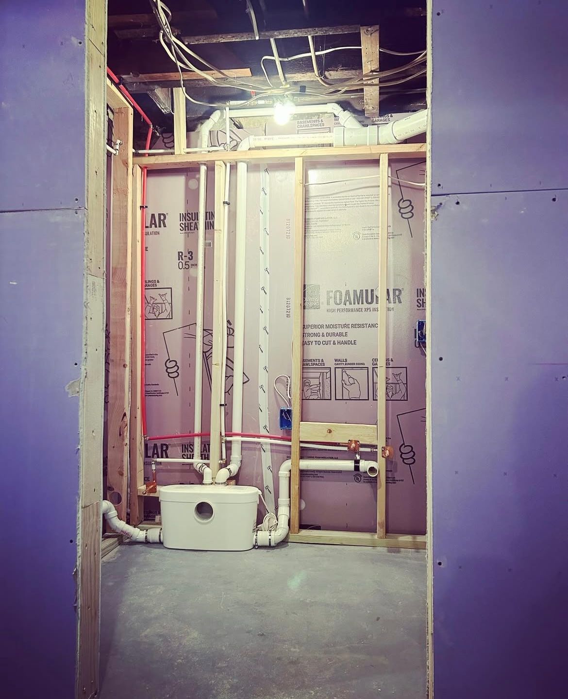 Interior of a bathroom under construction with framing, plumbing, and insulation visible. Walls are purple and gray.