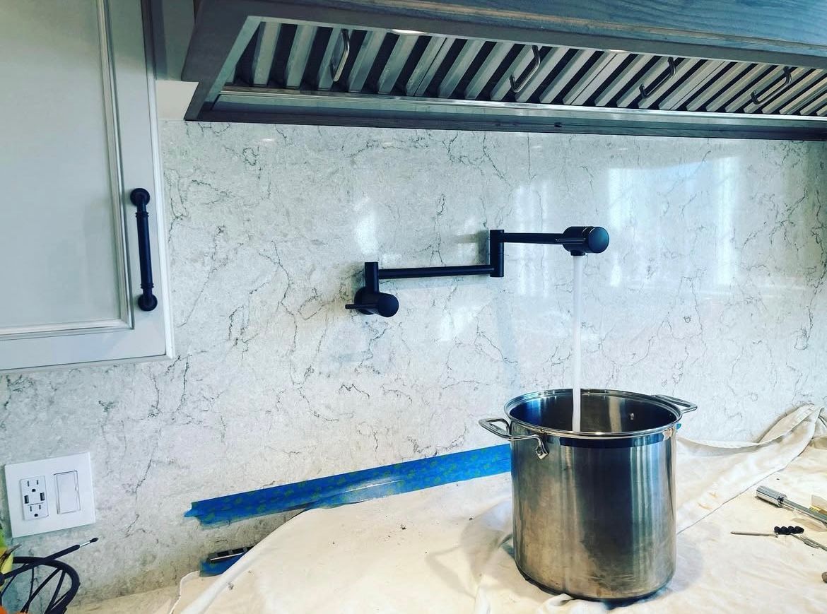 Pot filler faucet over a stainless steel pot on a kitchen countertop. Textured backsplash, range hood.