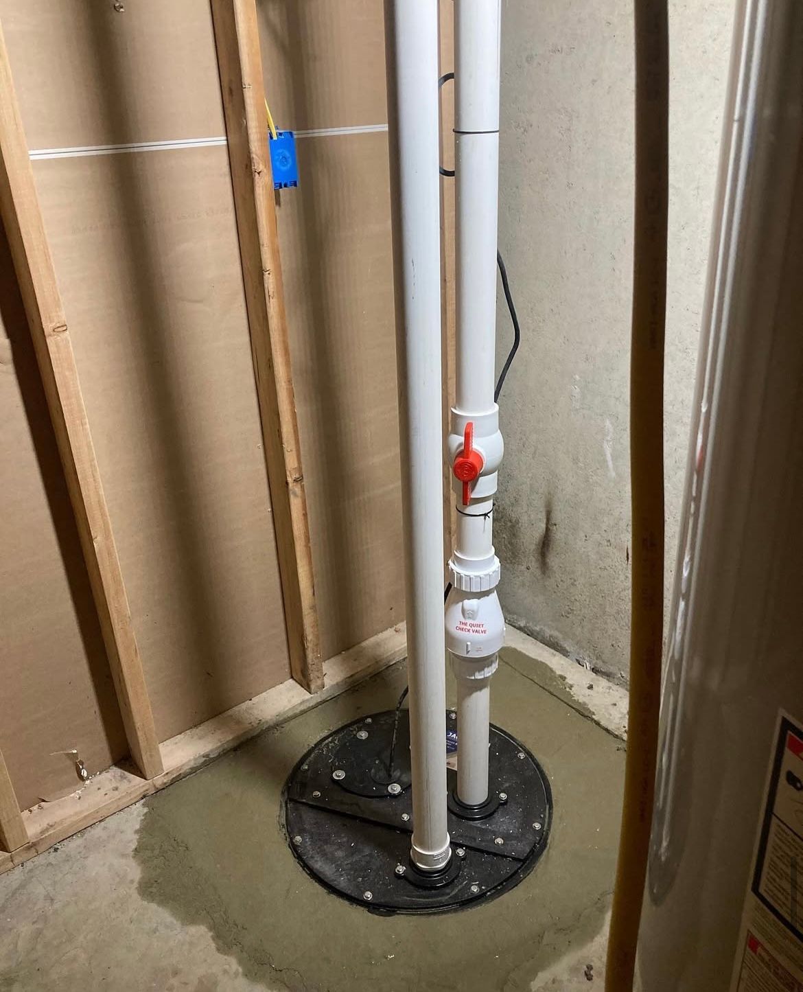Sump pump installation in a basement corner, with white pipes and a red valve, set in concrete.