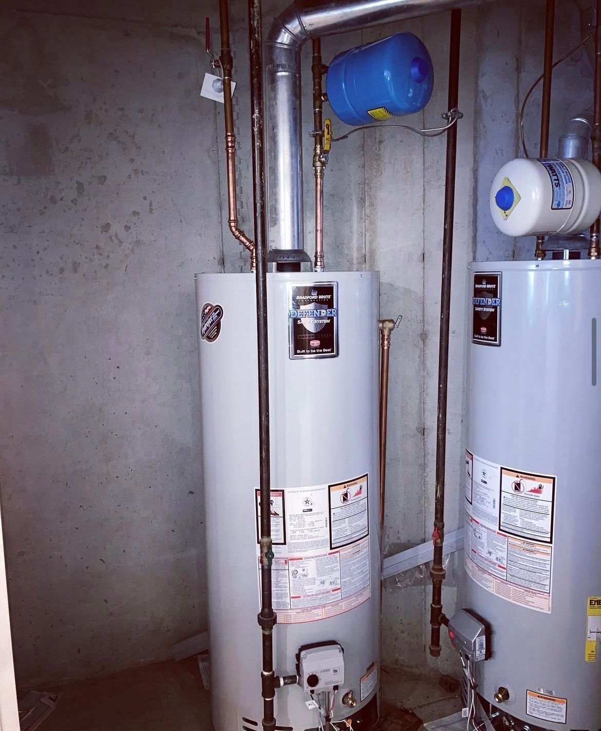 Two gray water heaters with copper pipes in a basement.