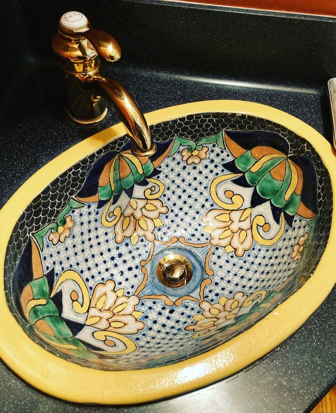 Oval, hand-painted ceramic sink with floral design and gold faucet on a dark countertop.