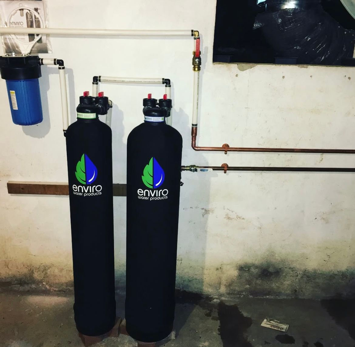 Water filtration system installed on a basement wall with black and blue filter tanks and white pipes.