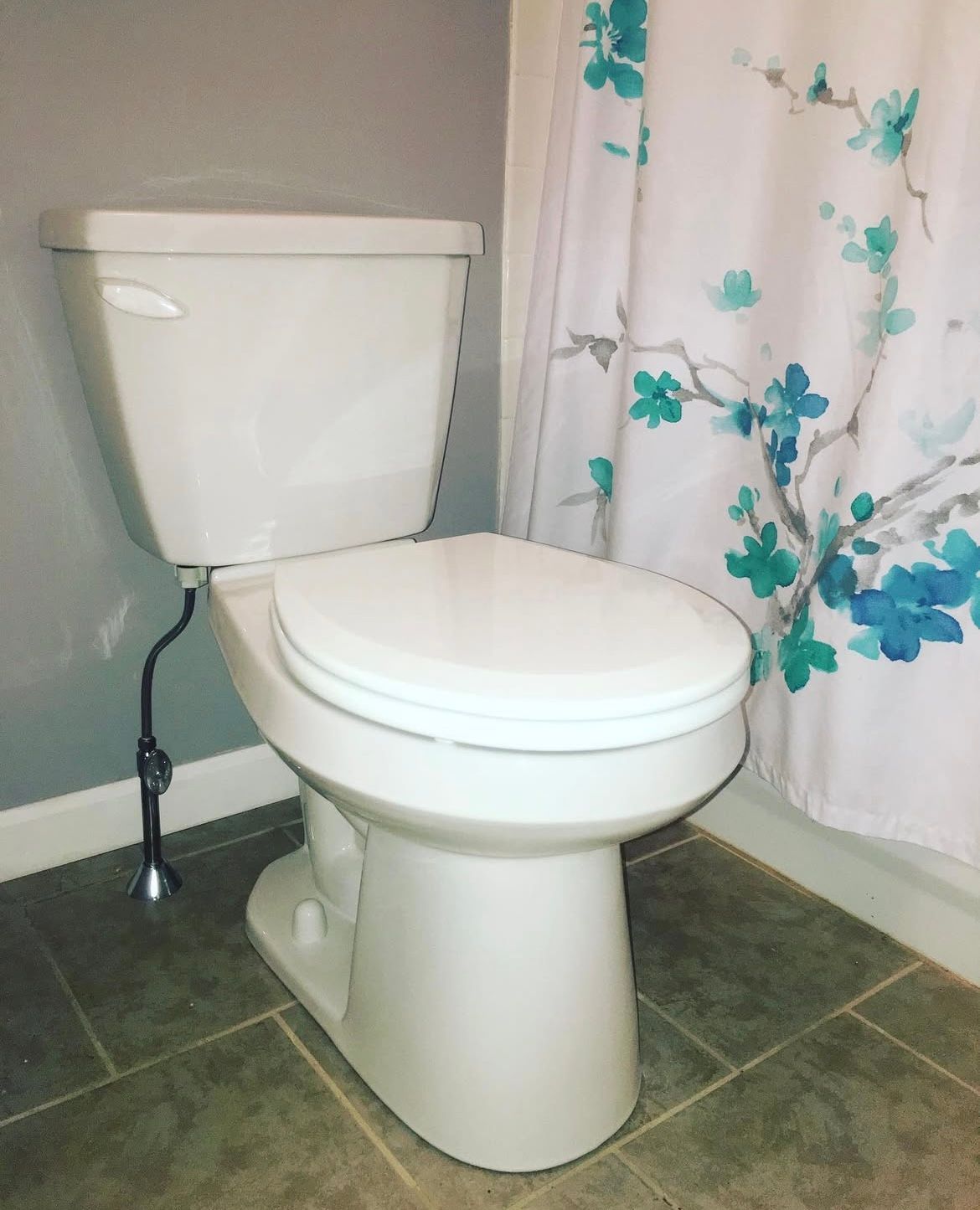 White toilet in a bathroom, with a floral shower curtain, against a gray wall and floor.