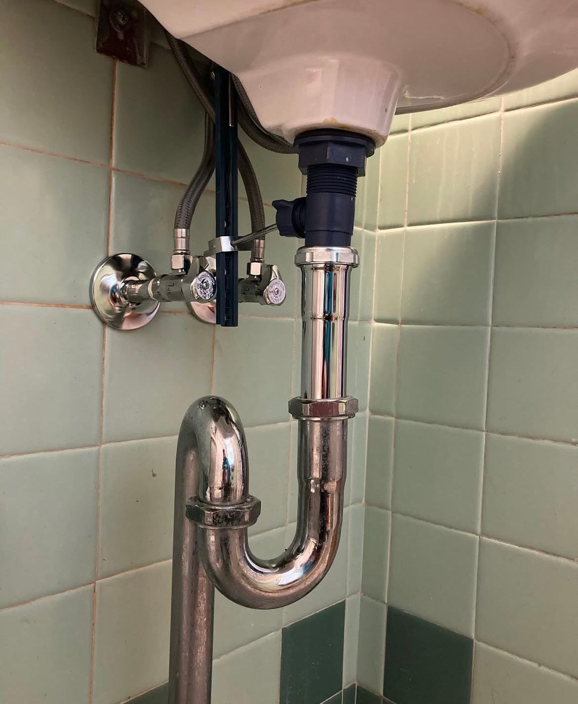 Plumbing under a white sink in a tiled bathroom. Chrome pipes and valves are visible.