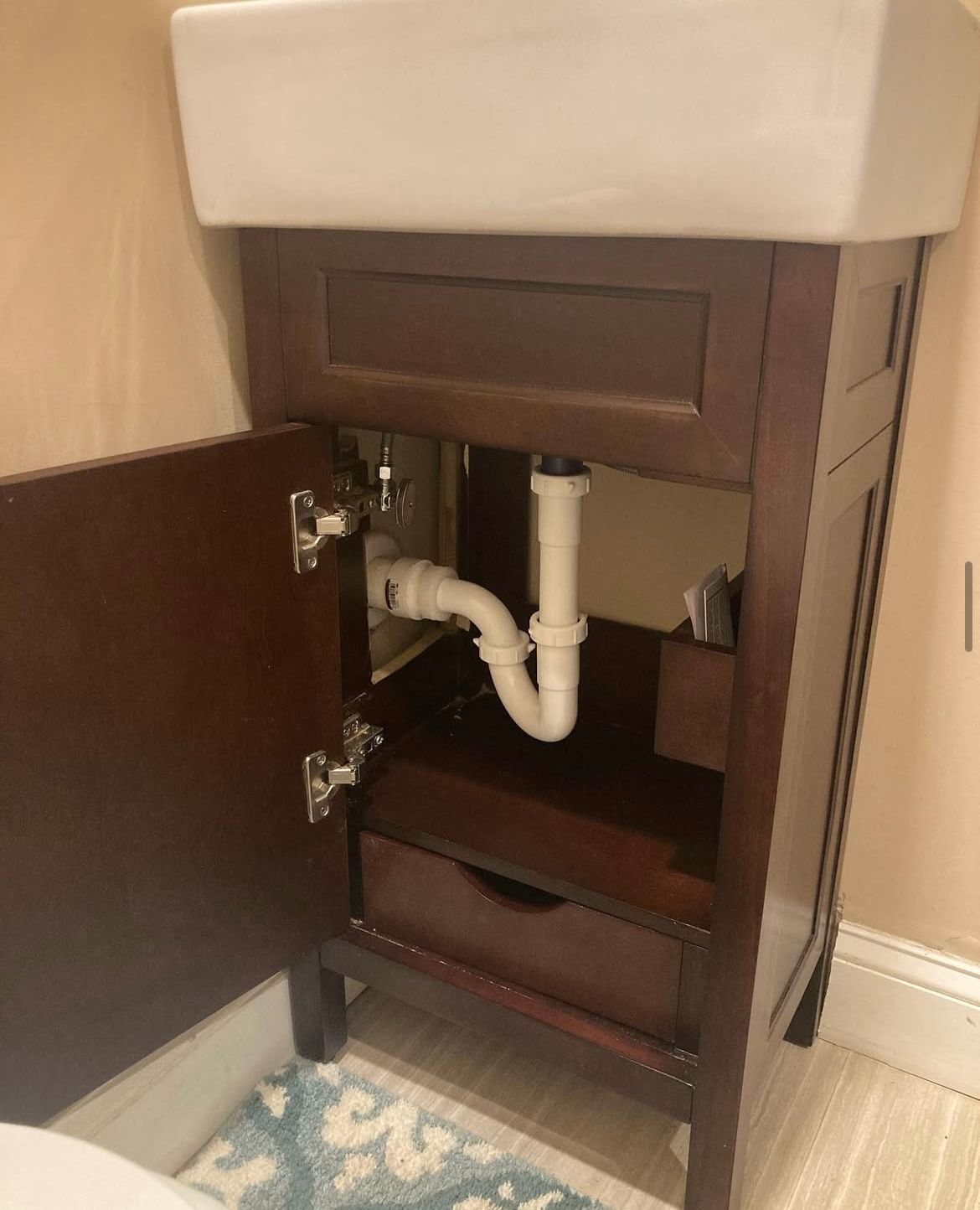 Bathroom vanity with open cabinet door, showing plumbing and drawer. Dark wood finish. White sink basin.