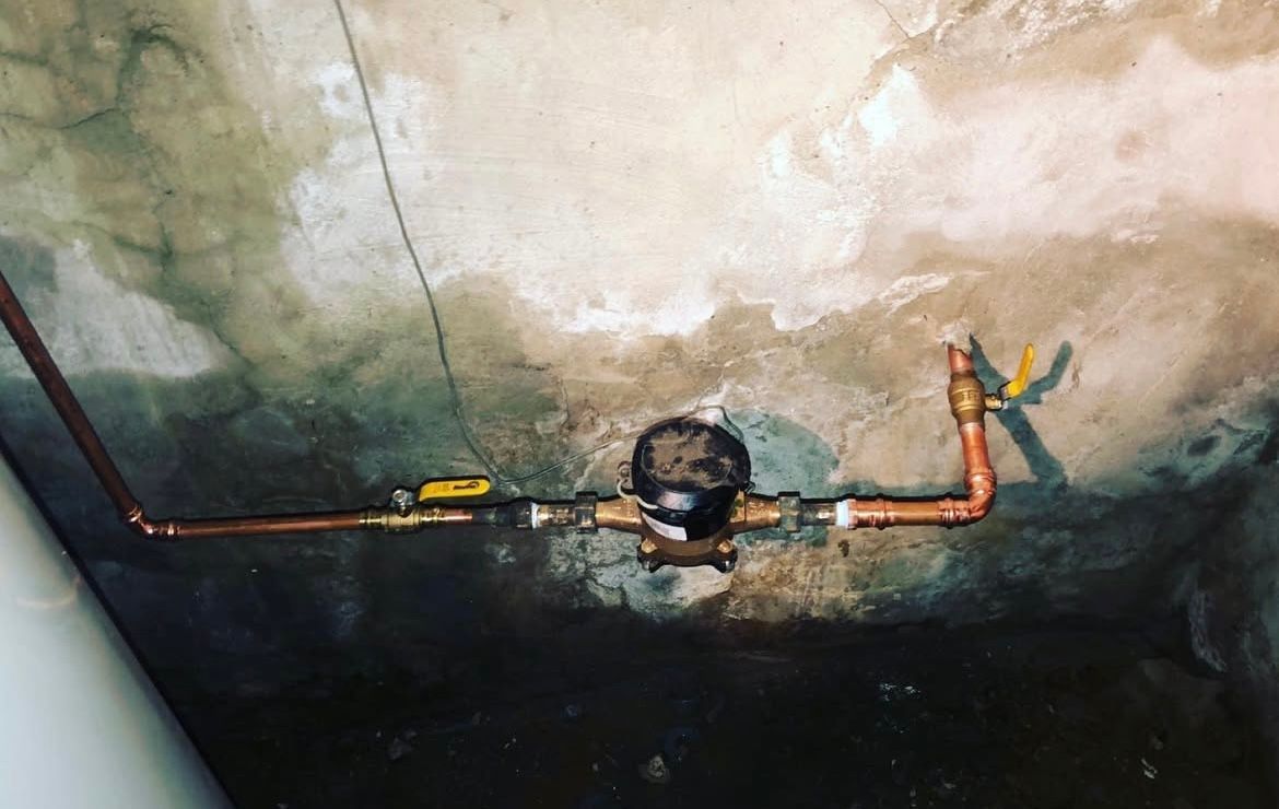 Copper pipes and valves against a weathered concrete wall. Water meter is visible.