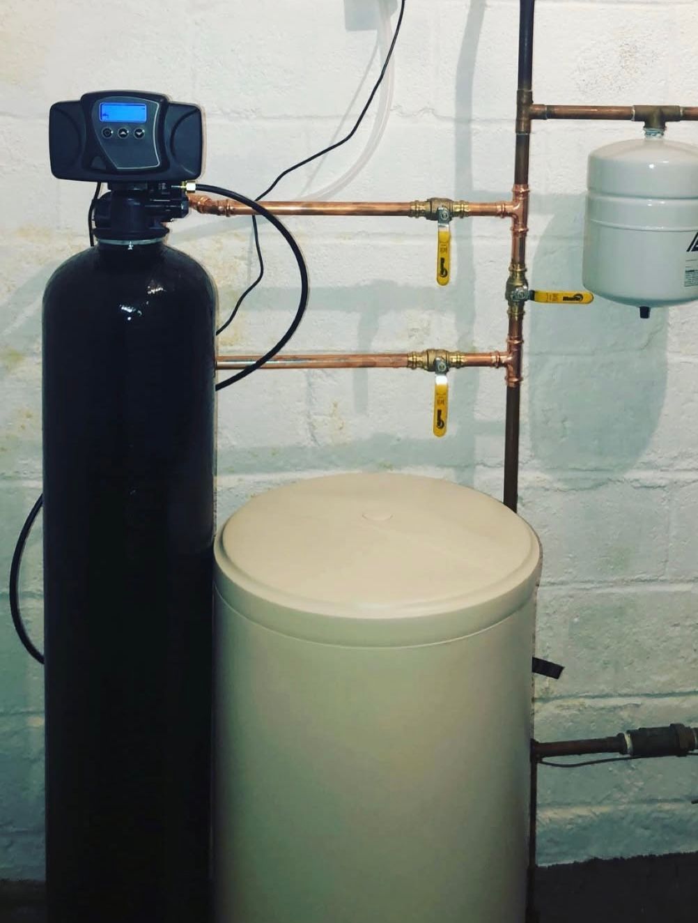 Water softener system in a basement setting with copper pipes, a black tank, and a white brine tank.