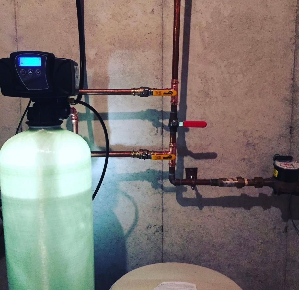 Water softener system with copper pipes, a digital display, and a red valve against a concrete wall.