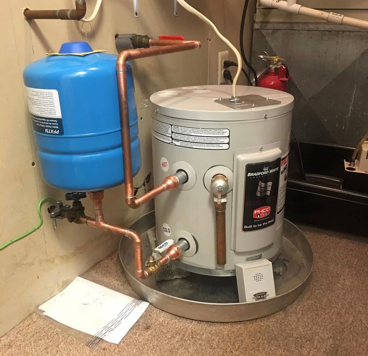 Blue expansion tank and copper-piped hot water heater with a catch pan in a utility room.