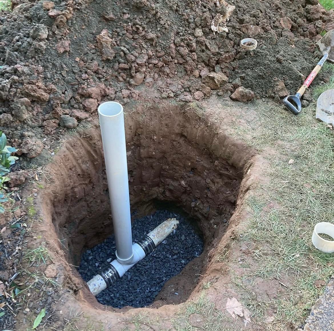 Hole dug in ground with PVC pipe and fittings installed. Gravel at bottom, dirt and shovel nearby.