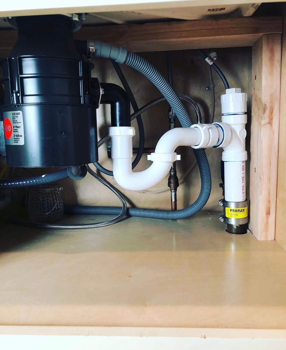 Undersink plumbing with garbage disposal. White pipes, gray hose, black disposal, wooden cabinet.