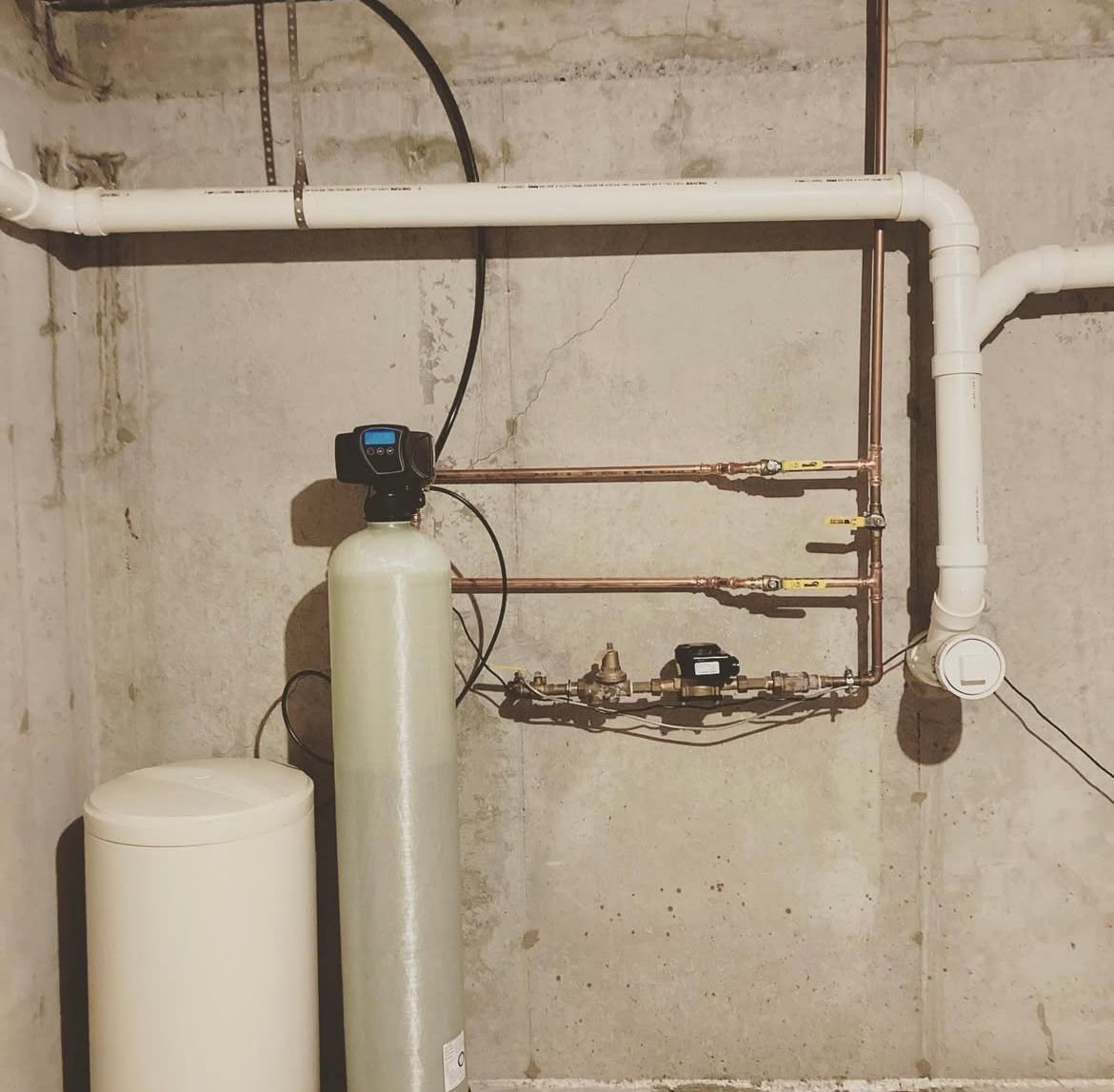 Water softener system in a basement with pipes, tanks, and valves against a concrete wall.