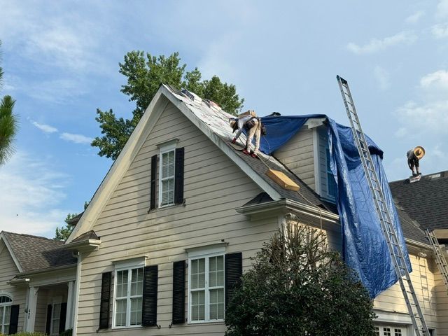 Roofers repairing a beige house roof with blue tarp and ladder under a clear sky