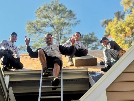 Four people sitting on a roof edge, smiling and waving against a blue sky and trees