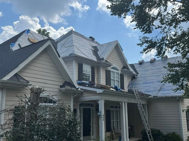 Two-story house with gray siding and roof, ladders leaning against the front and right side under a blue sky.