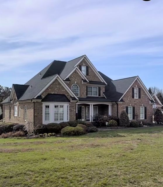 Large brick suburban house with dark roof and white trim, set on a grassy lawn under a blue sky