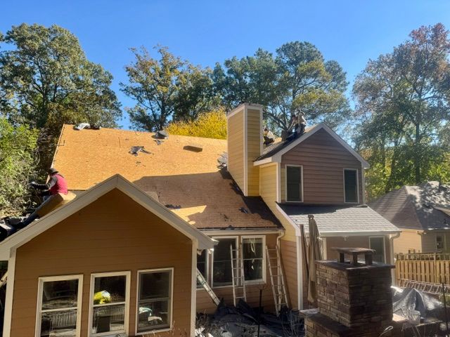House roof being repaired, with workers on top and a large chimney against a backdrop of trees.
