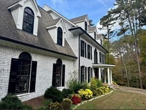 Large white two-story house with black shutters, porch, and landscaped garden by the woods