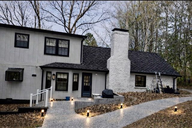 Two-story white house with black trim, lit walkway, and stone chimney in a wooded setting