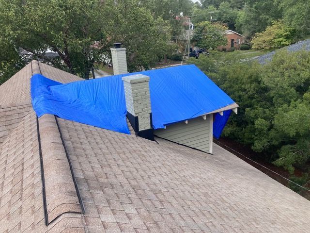Blue tarp covering part of a house roof amid trees and neighboring homes, seen from above