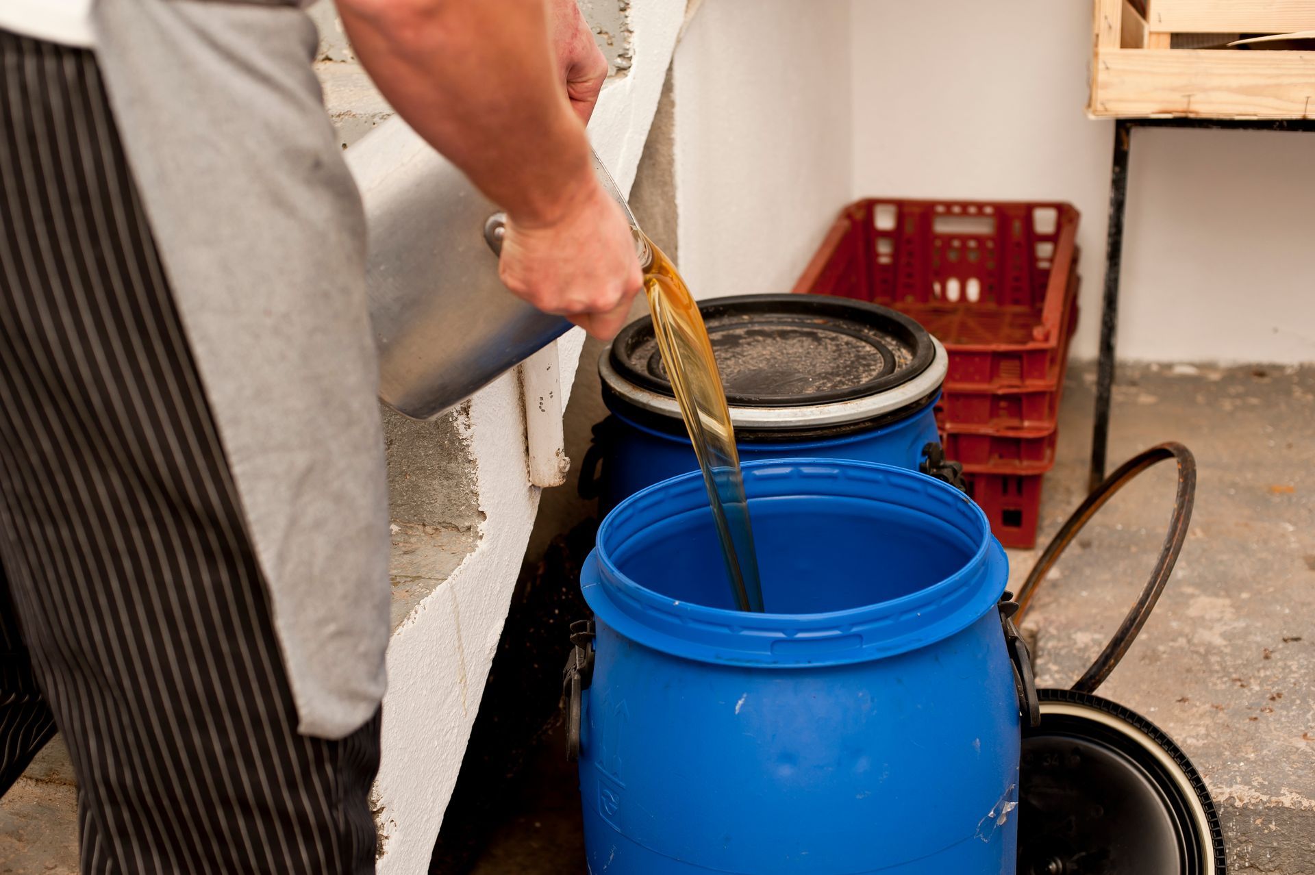 A man pouring used cooking oil from a metal container into a large blue plastic barrel