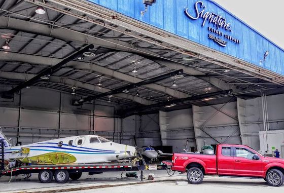 A red truck is parked in front of a hangar with a plane on a trailer.