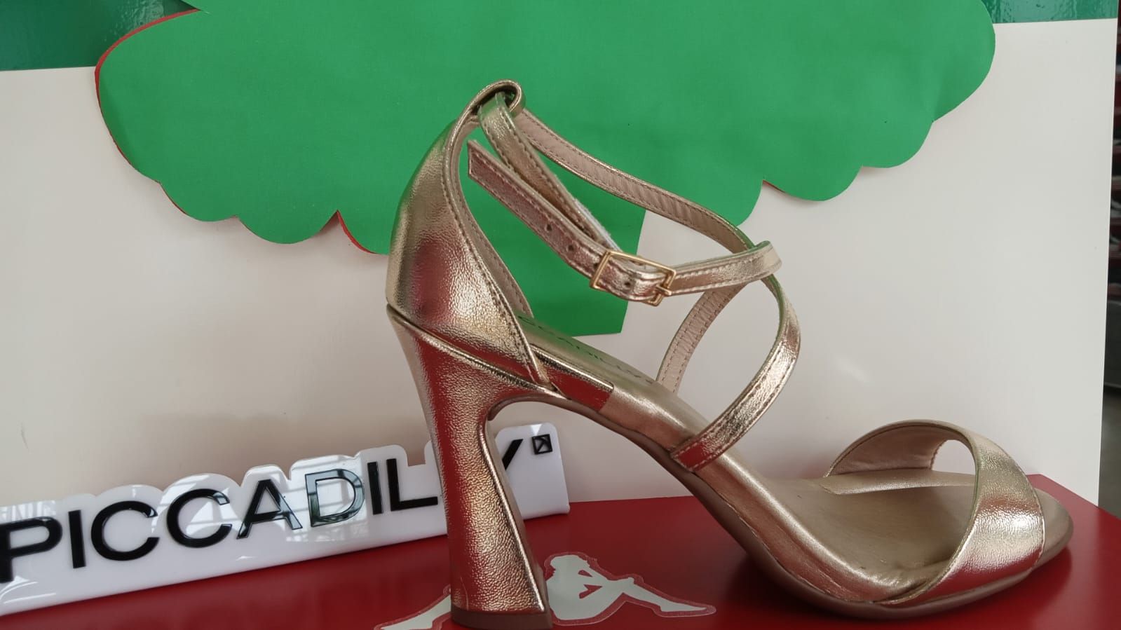 Sandalia de tacón alto de color dorado sobre superficie roja, con fondo blanco y verde.