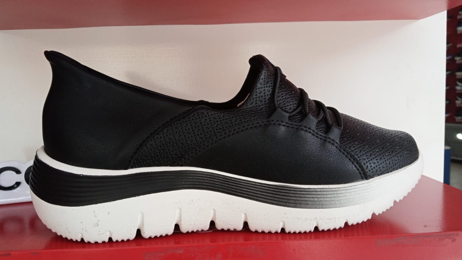 Zapatilla deportiva de mujer en color blanco y negro con suela gruesa blanca sobre un estante rojo.