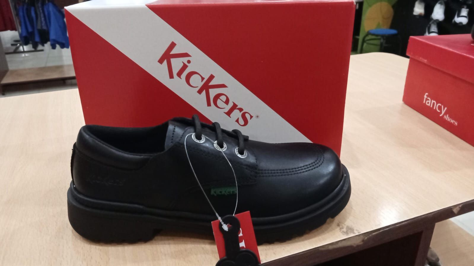 Zapato escolar Kickers negro con una caja Kickers roja sobre una mesa.